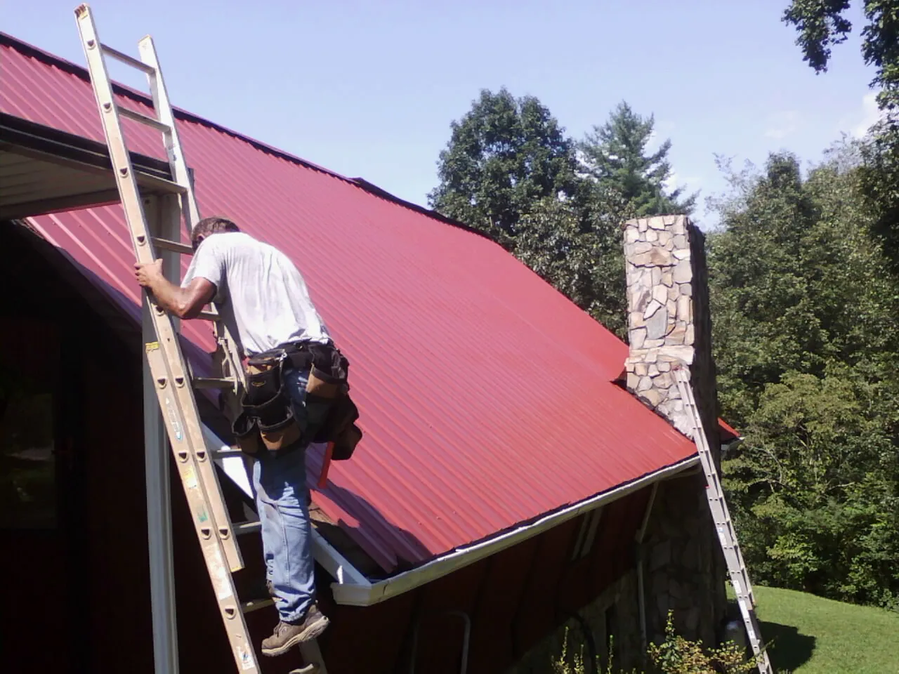 Roofer on a ladder beside a red metal roof and stone chimney — Soffit & Fascia Repair by Roofing Brothers in Cedar Falls