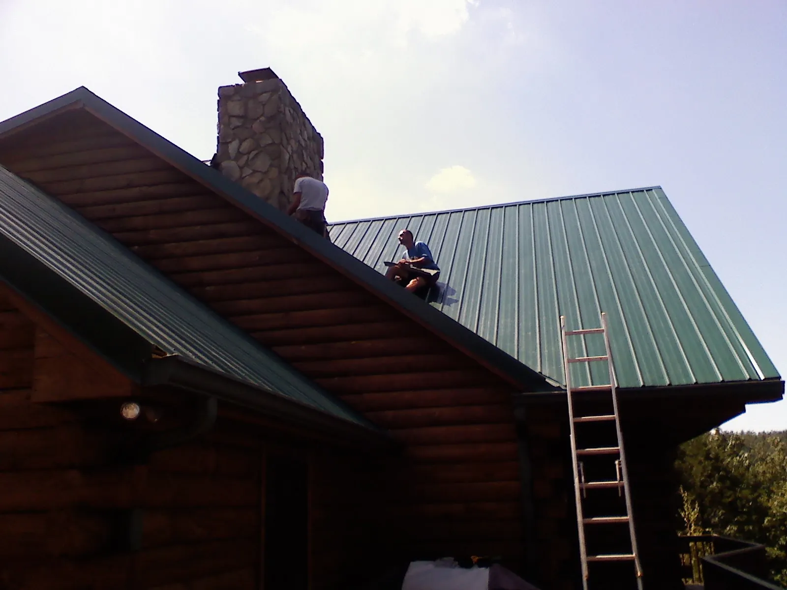 Roofers installing green metal panels beside a stone chimney on a log home in Cedar Falls